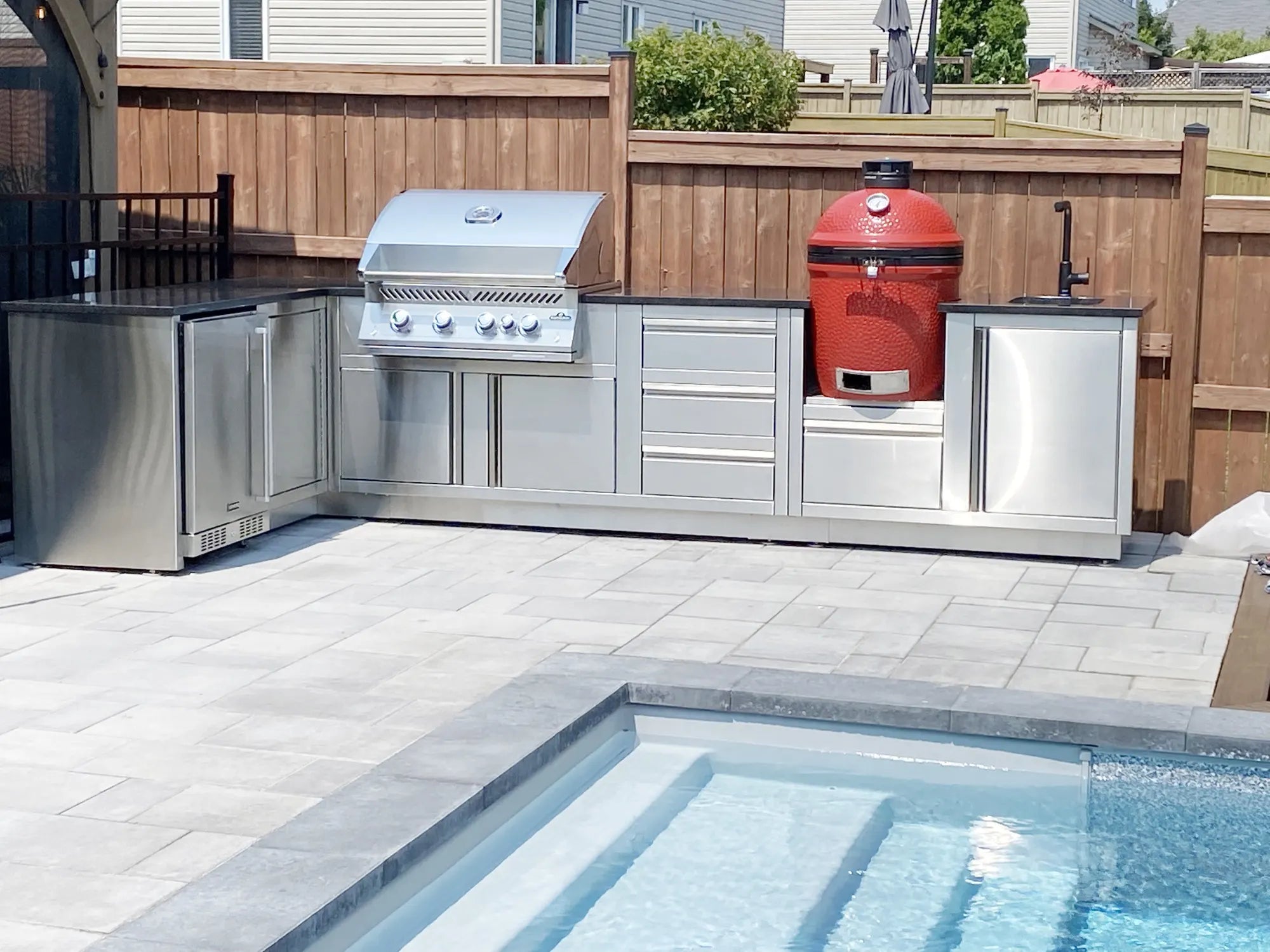 Cuisine extérieure Cuinox en L avec barbecue encastré et kamado rouge au bord de la piscine